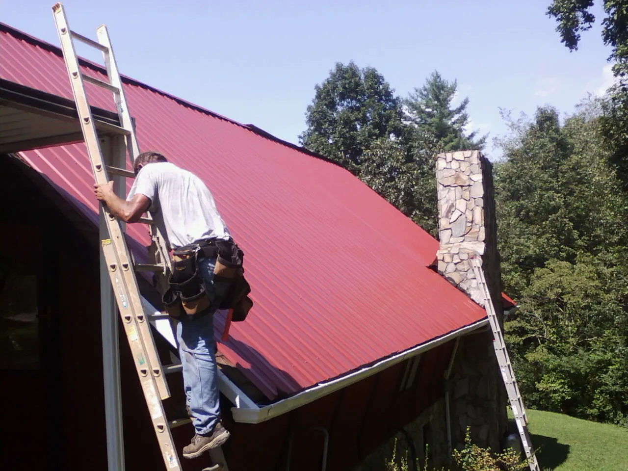 Roofer on a ladder beside a red metal roof and stone chimney — Soffit & Fascia Repair by Roofing Brothers in East Elmhurst